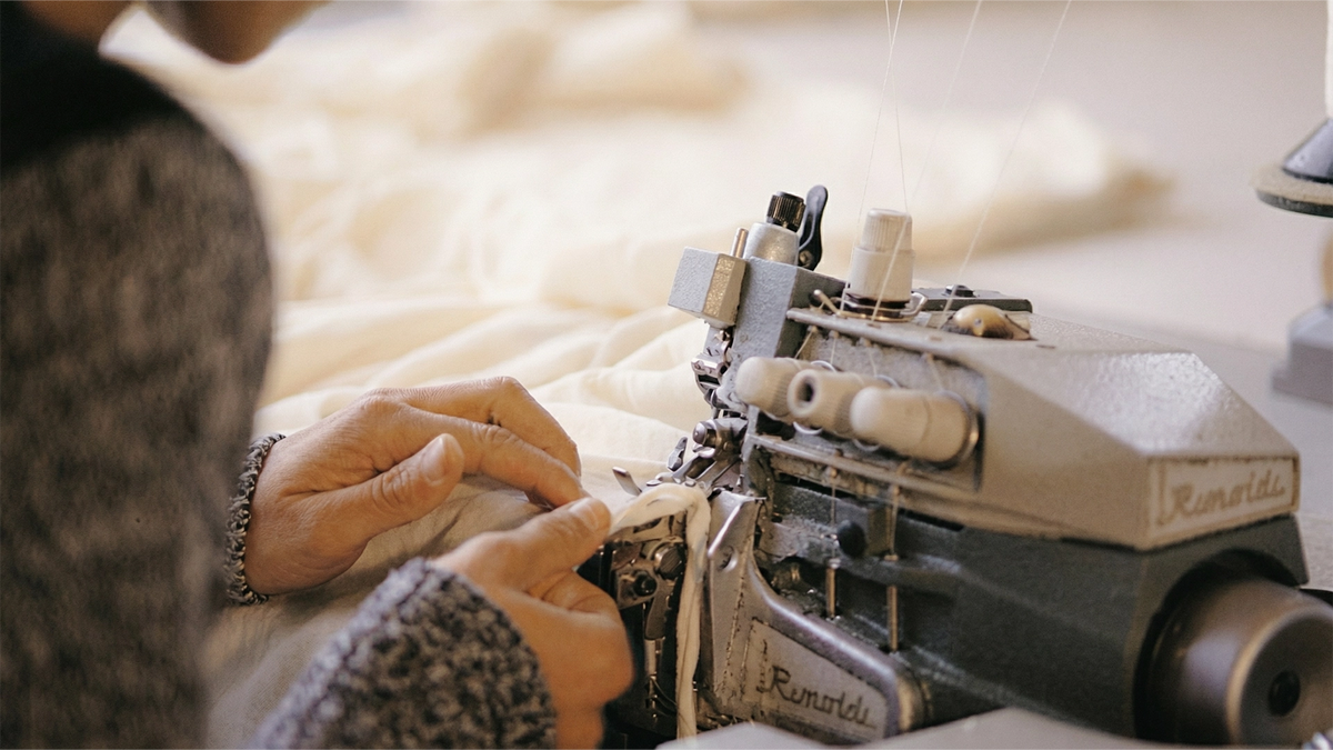 Nahaufnahme von Händen, die Stoff durch eine Industrienähmaschine führen, wobei unter der Nadel ein beiger Stoff zu sehen ist und rund um die Maschine Nähwerkzeuge zu erkennen sind.